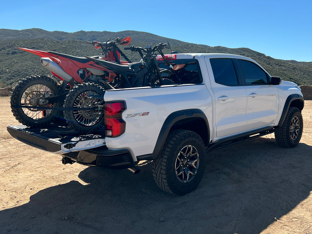 A Chevy Colorado truck with an ebike and a dirt bike motorcycle loaded into the bed on a desert landscape.