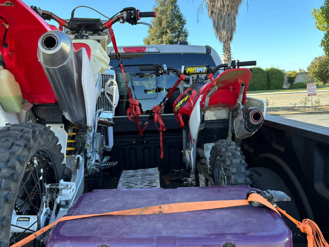 Two dirt bikes secured in the bed of a Ford Super Duty pickup using a CCR Sport truck bed moto rack system, viewed from inside the truck bed with tie-down straps and gear storage visible.