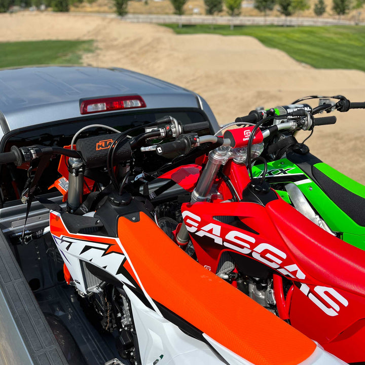 A colorful image of three dirt bikes loaded securely into the bed of a truck with the Hybrid Moto Rack three bike system. The back drop is a beautiful motocross race track.