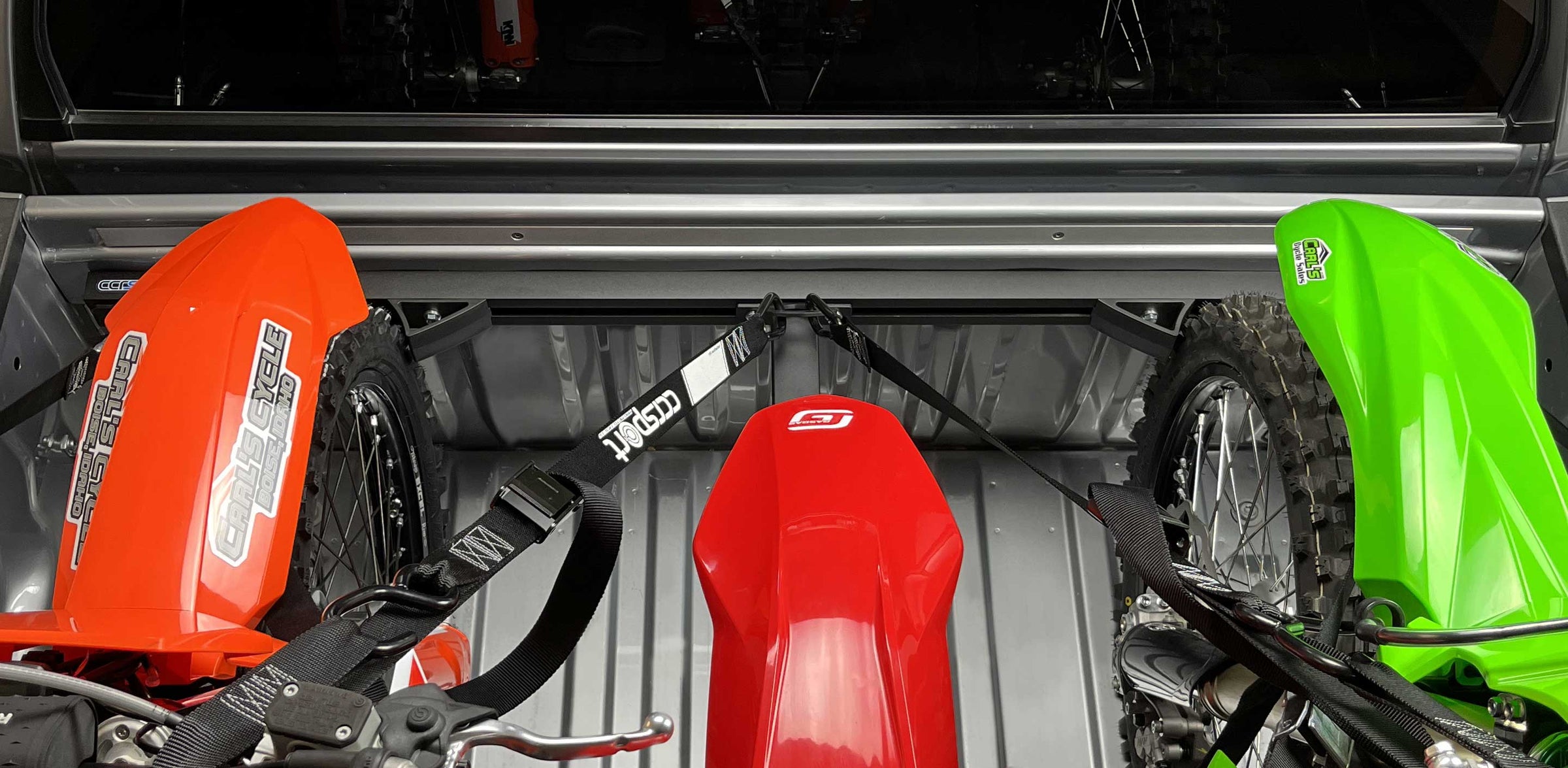 Three dirt bikes in a truck bed loaded with moto rack and offset wheel chock.