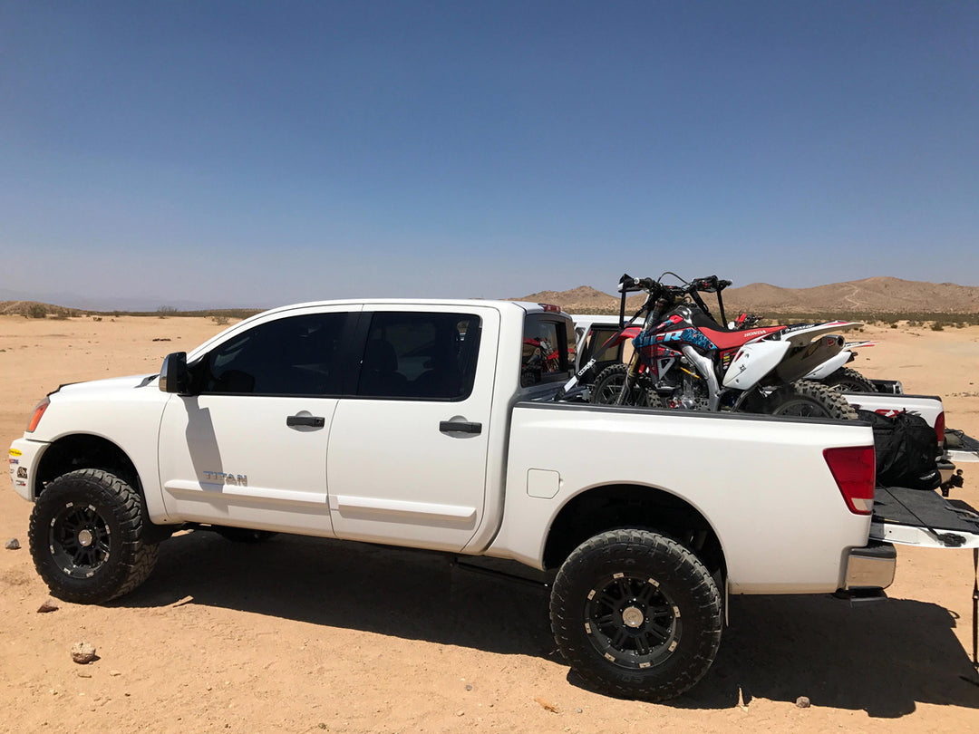 A white Nissan Titan with off-road tires and lift with a dirt bike loaded into the bed parked in the desert.
