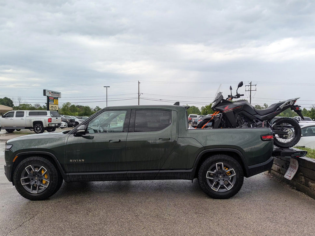 A forest green Rivian truck with an adventure motorcycle loaded in the trucks bed.