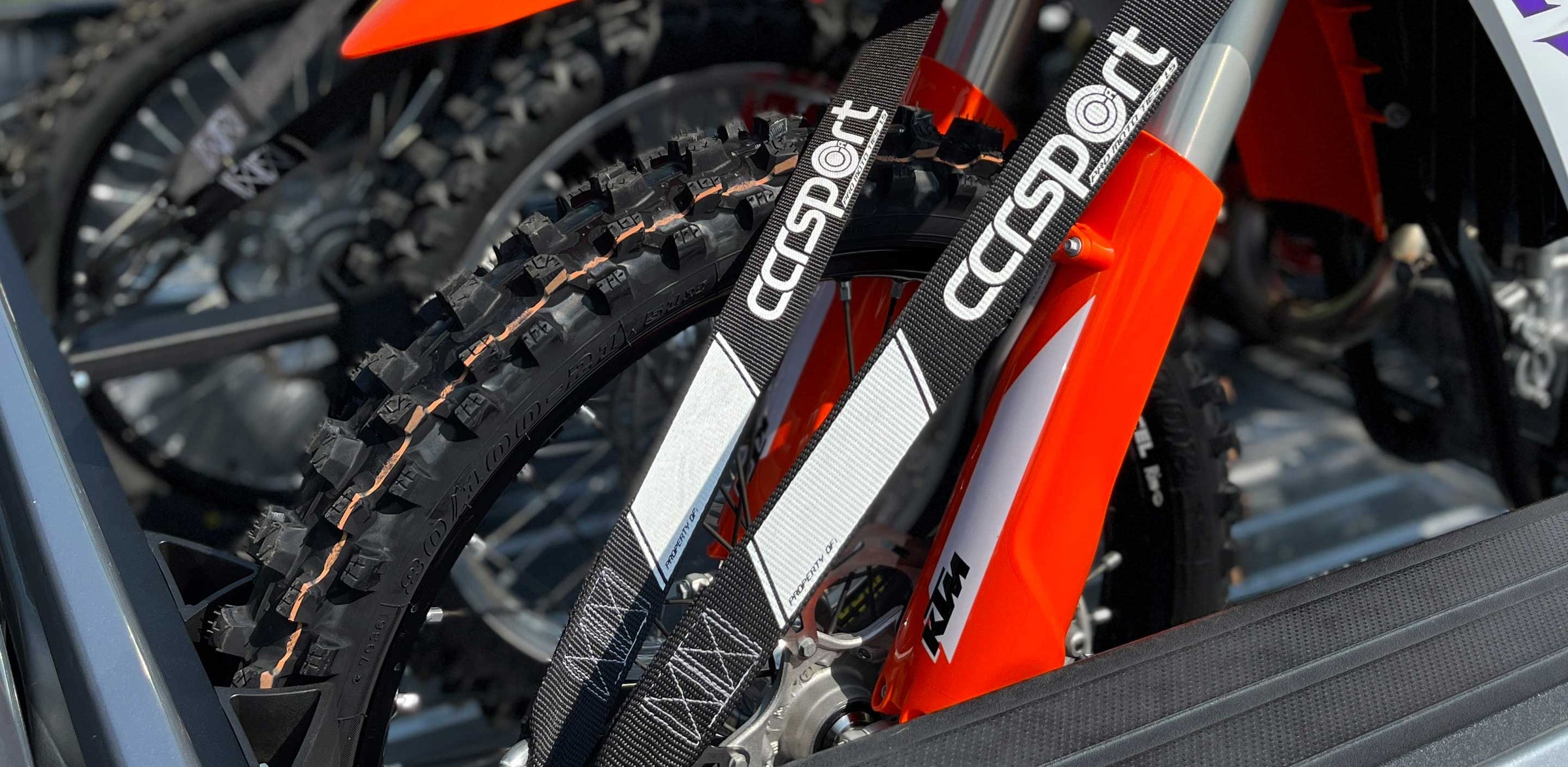 Three motorcycles loaded into a truck bed with detailed focus of a pair of tie down straps securing an orange KTM dirt bike.