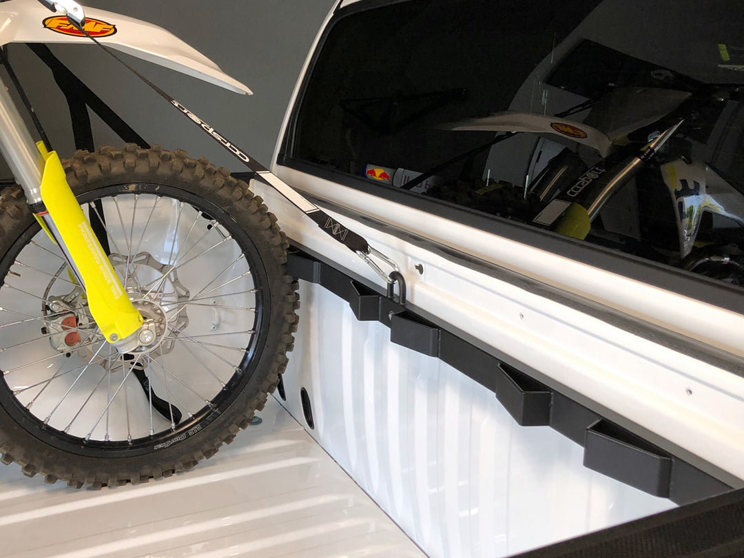 A nice studio photo of a white truck featuring the Bed Buddy motorcycle tie down rack with a dirt bike tied down in the trucks bed.