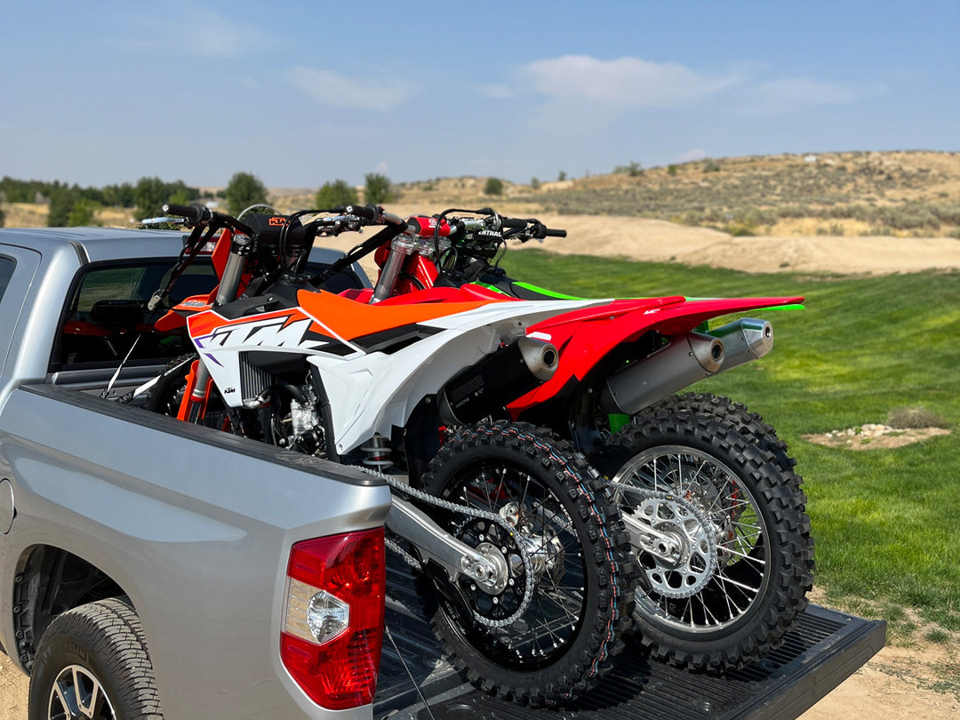 Three dirt bikes loaded into the bed of a truck parked on a scenic supercross track.