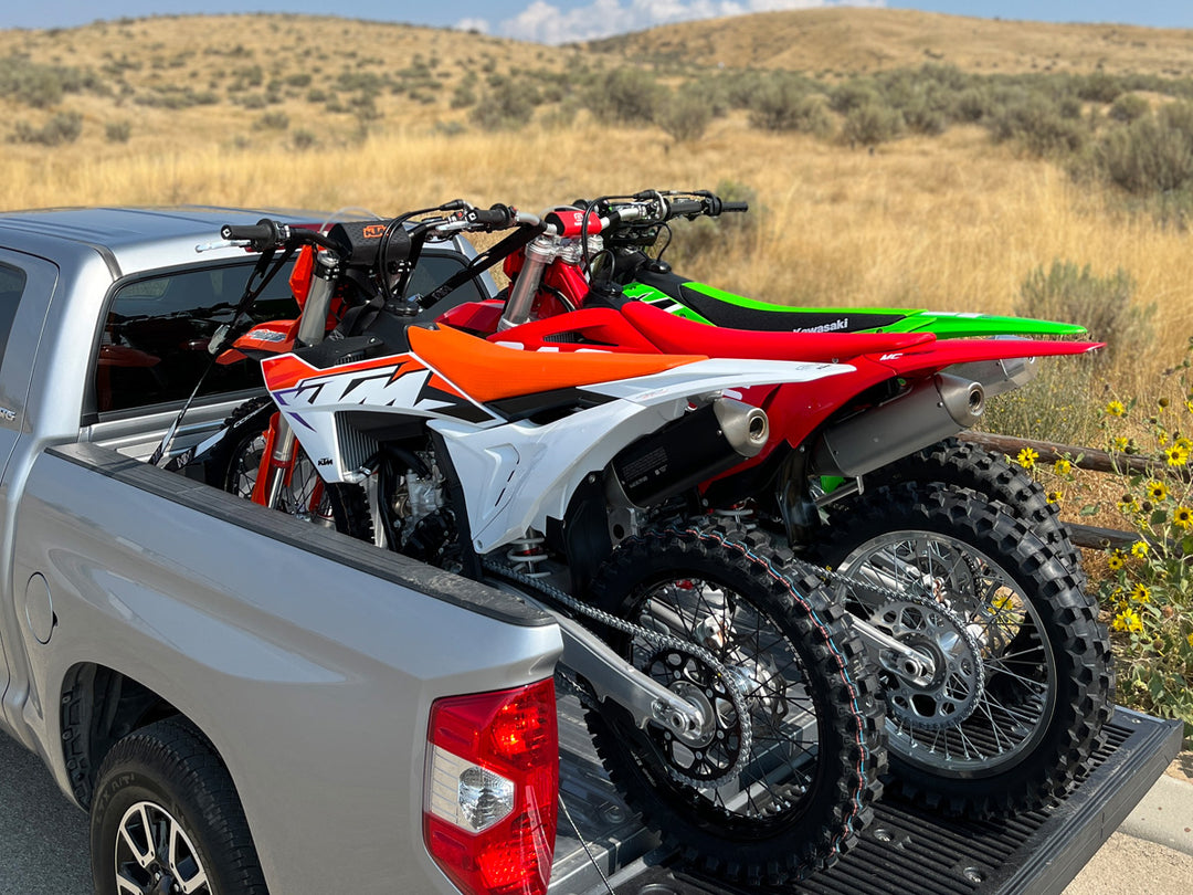 An orange, red and green dirt bikes loaded in a truck bed parked near grassy desert hills.